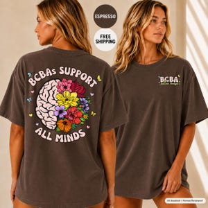 Puede incluir: Una camiseta de manga corta de color marrón oscuro con un gráfico en la espalda de un cerebro parcialmente compuesto por flores coloridas con el texto "BCBAs SUPPORT ALL MINDS" y un pequeño logo "BCBA Behavior Analyst" en el pecho.