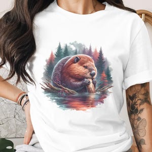 May include: White t-shirt featuring a detailed illustration of a beaver gnawing on a log in a serene forest setting. The artwork showcases the beaver in shades of brown and red, surrounded by trees and water.