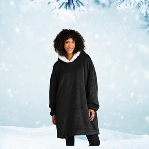 May include: A black oversized hoodie with a white fleece lining around the hood. The person is wearing the hoodie in a snowy winter scene. The sleeves are long, and the garment appears soft and comfortable. The hoodie is a warm winter garment.