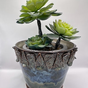May include: A ceramic planter with a blue and brown glaze, decorated with leaf-shaped accents. It holds three artificial succulents with green and yellow-green leaves. The planter is a decorative item for home decor.