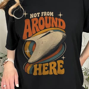 Könnte beinhalten: Ein schwarzes T-Shirt mit kurzen Ärmeln und einem Grafikdruck eines beigen Raumschiffs, das von bunten konzentrischen Ringen umgeben ist. Der Text "NOT FROM AROUND HERE" ist in einer orangefarbenen Retro-Schrift über und unter dem Motiv gedruckt.