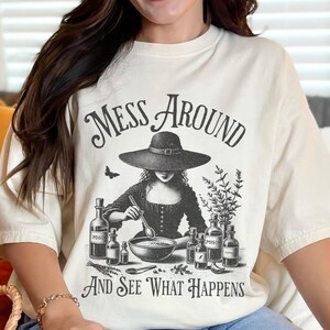Woman wearing off white Halloween Witch T-Shirt featuring Mess Around And See What Happens and a Witch Making Poison graphic. Perfect funny Halloween tee for a witchy aesthetic, spooky season, or a witch costume. Womens villain shirt for Halloween.