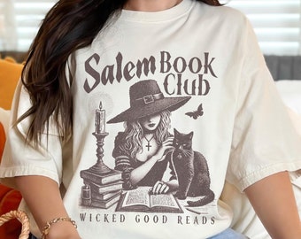 Könnte beinhalten: Elfenbeinfarbenes T-Shirt mit einem sepiafarbenen Aufdruck einer Hexe, einer schwarzen Katze und einer Kerze sowie dem Text "Salem Book Club" und "Wicked Good Reads". Das T-Shirt hat eine lockere Passform.