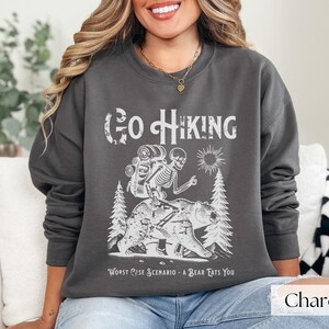 Woman wearing grey Go Hiking Worst Case Scenario A Bear Kills You, Funny Camping Sweater featuring a skeleton on a bear. Perfect for outdoor enthusiasts and camper gifts. Ideal for Halloween and summer vacation.