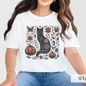 Woman wearing white Black Cat Halloween shirt featuring a spooky black cat, pumpkins, fall leaves, and flowers. Perfect for autumn and spooky season. Ideal for Halloween parties or as a cat lover gift with a vintage floral and witchy aesthetic.
