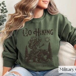 Woman wearing green Go Hiking Worst Case Scenario A Bear Kills You, Funny Camping Sweater featuring a skeleton on a bear. Perfect for outdoor enthusiasts and camper gifts. Ideal for Halloween and summer vacation.