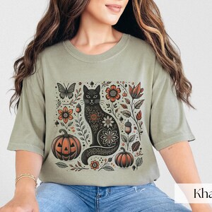 Woman wearing beige Black Cat Halloween shirt featuring a spooky black cat, pumpkins, fall leaves, and flowers. Perfect for autumn and spooky season. Ideal for Halloween parties or as a cat lover gift with a vintage floral and witchy aesthetic.