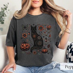 Woman wearing Black Cat Halloween shirt featuring a spooky black cat, pumpkins, fall leaves, and flowers. Perfect for autumn and spooky season. Ideal for Halloween parties or as a cat lover gift with a vintage floral and witchy aesthetic.