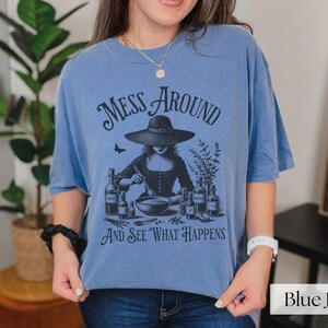 Woman wearing blue Halloween Witch T-Shirt featuring Mess Around And See What Happens and a Witch Making Poison graphic. Perfect funny Halloween tee for a witchy aesthetic, spooky season, or a witch costume. Womens villain shirt for Halloween.