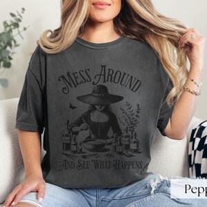 Woman wearing off black Halloween Witch T-Shirt featuring Mess Around And See What Happens and a Witch Making Poison graphic. Perfect funny Halloween tee for a witchy aesthetic, spooky season, or a witch costume. Womens villain shirt for Halloween.