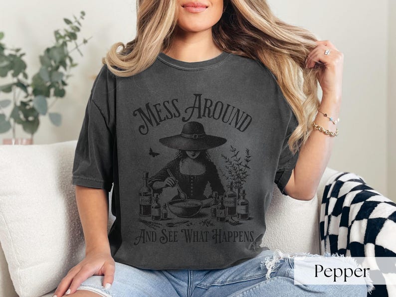 Woman wearing off black Halloween Witch T-Shirt featuring Mess Around And See What Happens and a Witch Making Poison graphic. Perfect funny Halloween tee for a witchy aesthetic, spooky season, or a witch costume. Womens villain shirt for Halloween.