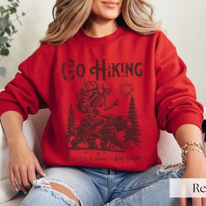 Woman wearing red Go Hiking Worst Case Scenario A Bear Kills You, Funny Camping Sweater featuring a skeleton on a bear. Perfect for outdoor enthusiasts and camper gifts. Ideal for Halloween and summer vacation.