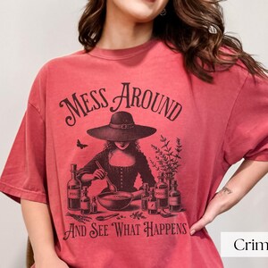 Woman wearing red Halloween Witch T-Shirt featuring Mess Around And See What Happens and a Witch Making Poison graphic. Perfect funny Halloween tee for a witchy aesthetic, spooky season, or a witch costume. Womens villain shirt for Halloween.