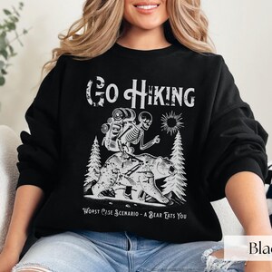 Woman wearing black Go Hiking Worst Case Scenario A Bear Kills You, Funny Camping Sweater featuring a skeleton on a bear. Perfect for outdoor enthusiasts and camper gifts. Ideal for Halloween and summer vacation.