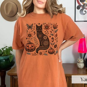 Woman wearing orange Black Cat Halloween shirt featuring a spooky black cat, pumpkins, fall leaves, and flowers. Perfect for autumn and spooky season. Ideal for Halloween parties or as a cat lover gift with a vintage floral and witchy aesthetic.
