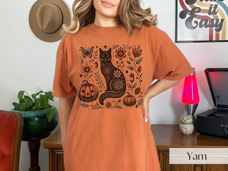 Woman wearing orange Black Cat Halloween shirt featuring a spooky black cat, pumpkins, fall leaves, and flowers. Perfect for autumn and spooky season. Ideal for Halloween parties or as a cat lover gift with a vintage floral and witchy aesthetic.