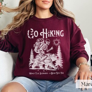 Woman wearing maroon Go Hiking Worst Case Scenario A Bear Kills You, Funny Camping Sweater featuring a skeleton on a bear. Perfect for outdoor enthusiasts and camper gifts. Ideal for Halloween and summer vacation.