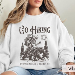 Woman wearing white Go Hiking Worst Case Scenario A Bear Kills You, Funny Camping Sweater featuring a skeleton on a bear. Perfect for outdoor enthusiasts and camper gifts. Ideal for Halloween and summer vacation.