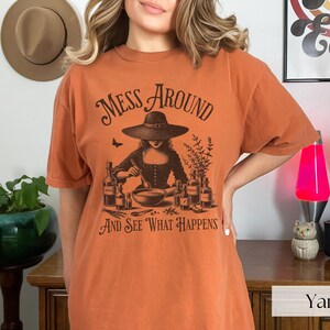 Woman wearing orange Halloween Witch T-Shirt featuring Mess Around And See What Happens and a Witch Making Poison graphic. Perfect funny Halloween tee for a witchy aesthetic, spooky season, or a witch costume. Womens villain shirt for Halloween.
