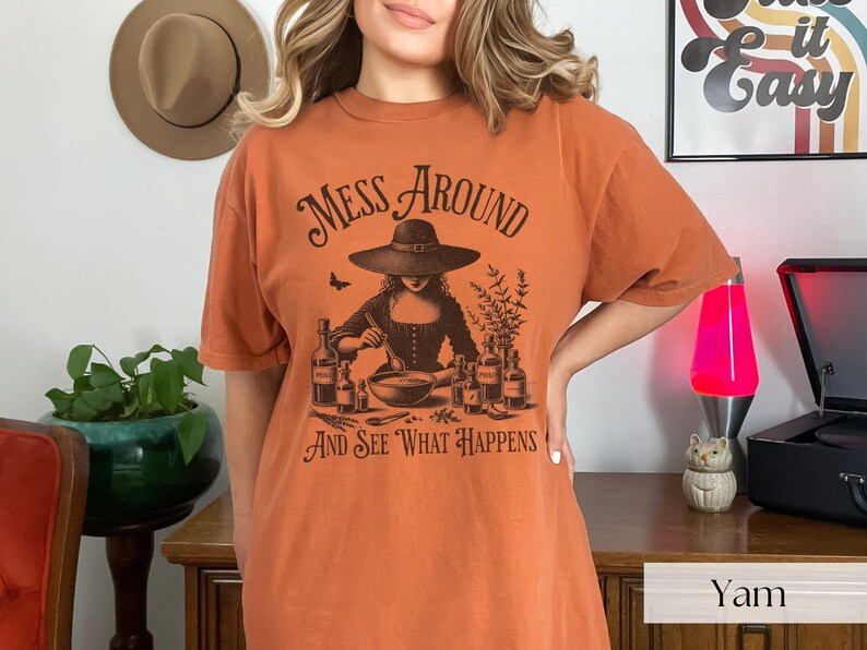 Woman wearing orange Halloween Witch T-Shirt featuring Mess Around And See What Happens and a Witch Making Poison graphic. Perfect funny Halloween tee for a witchy aesthetic, spooky season, or a witch costume. Womens villain shirt for Halloween.