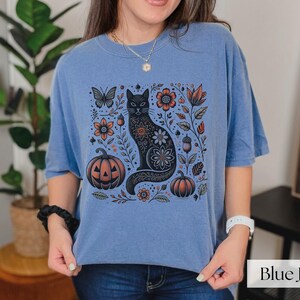 Woman wearing blue Black Cat Halloween shirt featuring a spooky black cat, pumpkins, fall leaves, and flowers. Perfect for autumn and spooky season. Ideal for Halloween parties or as a cat lover gift with a vintage floral and witchy aesthetic.