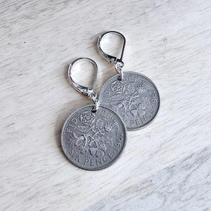 May include: Silver-toned leverback earrings featuring vintage Six Pence coins from 1967. The coins display floral and botanical designs, with the text "FID DEF" and "SIX PENCE". The earrings are displayed on a light wood grain surface.