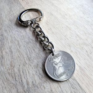 May include: A silver keychain with a 50-øre coin from Norway, dated 1964. The coin features a stylized image of a dog.