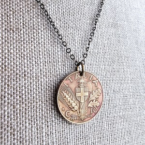 May include: A bronze-colored pendant necklace featuring a vintage Italian coin. The coin displays the word "ITALIA", a cross, wheat, and the year 1941. The chain is a dark, gunmetal color, and the pendant is round.