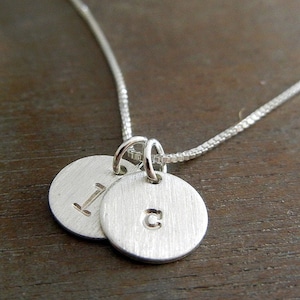 May include: A silver necklace featuring two circular pendants with the letters "I" and "C" engraved on them. The pendants are connected to a delicate chain, creating a personalized and elegant piece of jewelry.