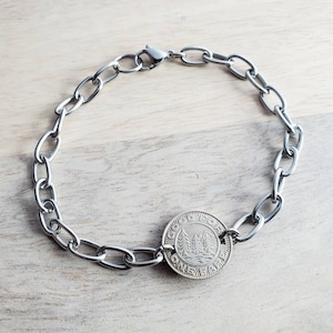 May include: Silver-coloured chain bracelet with an oval link design. The bracelet features a circular coin with a landscape design of trees and the text "ONE FARE". The bracelet is displayed on a light wood surface.