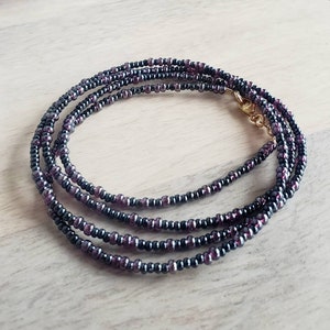 May include: A multi-strand beaded necklace with small, dark purple and black beads. The necklace is made of multiple strands of beads that are woven together.