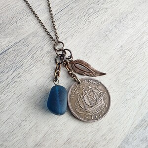 May include: A bronze-toned necklace featuring a blue sea glass bead, a silver coin with a ship design, and a bronze feather charm. The coin is dated 1964. The necklace has a delicate chain.