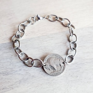 May include: A silver chain bracelet with a silver buffalo nickel charm. The nickel features the words "United States of America" and "Five Cents".