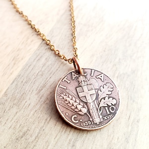 May include: A copper-colored coin pendant necklace with a gold-colored chain. The coin features the word "ITALIA" and the year 1939. The coin has a detailed design with wheat, a shield, and other symbols. The necklace is displayed on a light wood surface.