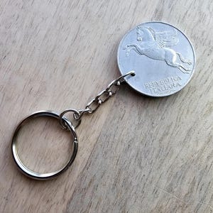 May include: A silver-colored keychain featuring a coin with a winged horse design. The coin is attached to a chain and a keyring. The text "REPUBBLICA ITALIANA" is visible on the coin. The background is a light brown wooden surface.