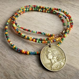 Könnte beinhalten: Eine mehrreihige Halskette aus kleinen, bunten Perlen in Blautönen, Grün, Orange und Gelb. Die Halskette hat einen Anhänger in Form einer runden Münze mit der Aufschrift "REPUBLIQUE FRANCAISE" und dem Profil einer Frau.