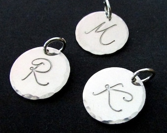 Peut inclure: Trois breloques en argent avec une finition martelée, chacune présentant une lettre différente en écriture cursive. Les lettres sont M, R et T.