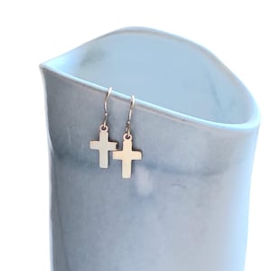 Może przedstawiać: Para złotych kolczyków w kształcie krzyża wiszących przed jasnoniebieskim ceramicznym wazonem. Kolczyki mają prosty design z zapięciem na haczyk. Wazon ma gładkie, matowe wykończenie.