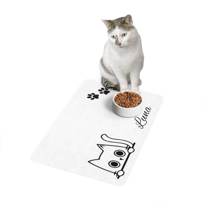 Peut inclure: Un chat blanc est assis pr&egrave;s d'un tapis blanc pour nourriture pour animaux de compagnie avec une illustration de chat noir et des empreintes de pattes. Un bol blanc rempli de nourriture pour chats est pos&eacute; sur le tapis. Le nom "Luna" est &eacute;crit sur le tapis.