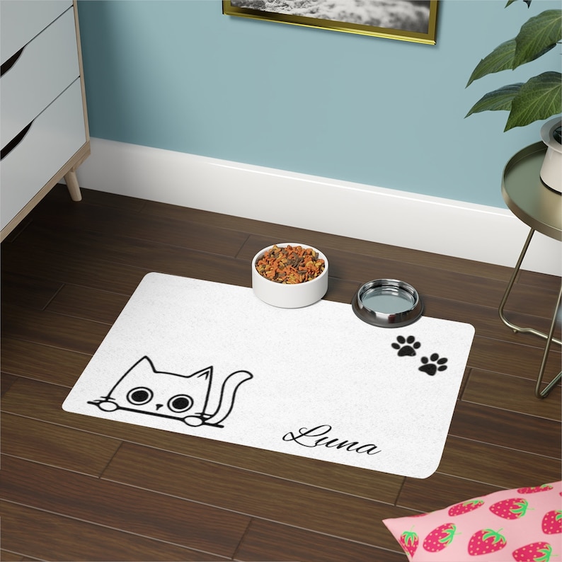 Peut inclure: Tapis de nourriture pour animaux de compagnie blanc avec un dessin de chat noir, le nom "Luna" et des empreintes de pattes. Comprend un bol en c&eacute;ramique blanc et un bol en acier inoxydable. Le tapis est sur un sol en bois fonc&eacute;.