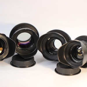 May include: Four black camera lenses with lens hoods and caps are arranged on a white surface. The lenses have a circular design with a ridged focus ring. The lenses are of varying sizes and have a reflective interior.