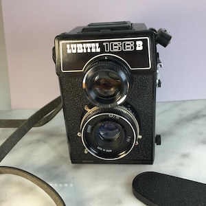 May include: Black Lubitel 166B twin-lens reflex camera with a leather strap. The camera has two lenses, one above the other, with the text "LUBITEL 166B" in white on the front. The lenses have silver rims and are labeled with numbers.