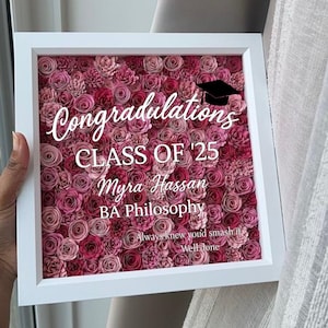 May include: A square white shadow box frame filled with numerous small paper flowers in various shades of pink and deep red. The glass front features white text reading Congratulations, CLASS OF '25, Myra Hassan, BA Philosophy, Always knew you'd smash it, Well done, and a black graduation cap icon.
