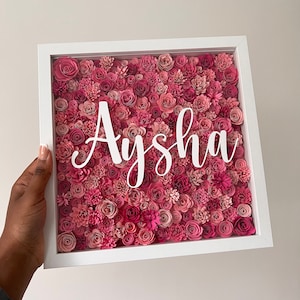 May include: A white framed floral artwork featuring an array of pink paper flowers. The name "Aysha" is written in white cursive across the center. The frame is held by a person's hand, showcasing the piece against a neutral background.