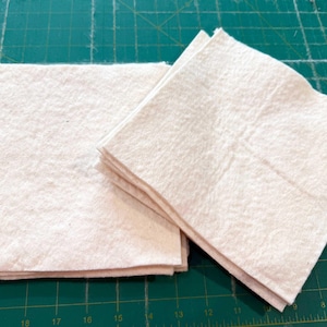 Puede incluir: Dos pilas de cuadrados de tela blanca, probablemente para proyectos de acolchado o manualidades.