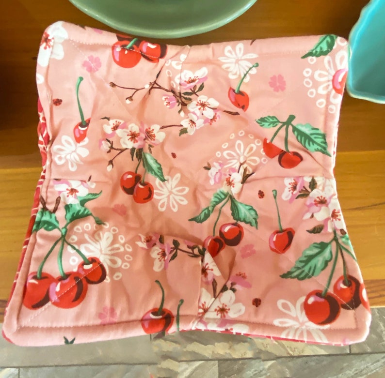 May include: A pink quilted hot pad with a cherry blossom and cherry print. The hot pad is square and has a red and white checkered border.