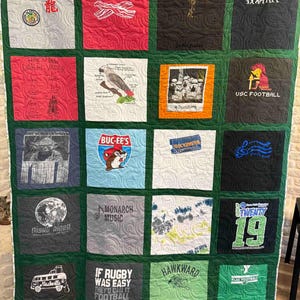 May include: A memory quilt composed of twenty square fabric panels featuring various graphic prints, including text, logos, and illustrations. The panels are stitched together with green fabric borders and finished with a swirling quilted pattern throughout.