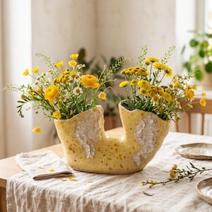 May include: A yellow ceramic vase with a dual-opening, organic shape and speckled finish. The vase features textured, white, leaf-like relief patterns on the front and is filled with a vibrant arrangement of yellow flowers and green stems.