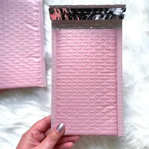 Puede incluir: Dos sobres de burbujas rosas con un sello plateado y negro estampado. Los sobres están hechos de un material suave y flexible y tienen un interior acolchado para proteger el contenido.