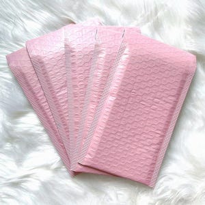 May include: Five pink bubble mailers, also known as padded envelopes, are stacked on top of each other. The mailers are made of a lightweight, flexible material with a textured surface.